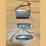 Tři elektrické snímače kytary s kovovými kryty v stříbrné barvě, umístěné na dřevěném povrchu. Horní snímač je typu humbucker s šesti viditelnými pol…