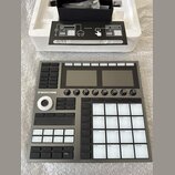 Šedý sampler Maschine+ s dvěma displeji, několika ovládacími knoflíky a bílými podsvícenými pady. Na levé straně je vidět nápis MASCHINE+ a ovládací…