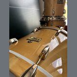 Detailní pohled na světlý, žlutohnědý lakovaný basový buben s kovovým kováním. Na korpusu je viditelný nápis Pearl Masters Custom SE. Povrch laku je…