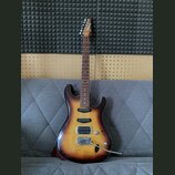 Elektrická kytara s trupem v barevně sunburst a hnedým hmatníkem. Na hlavě je vidět nápis Ibanez. Nástroj má dva humbuckery, jeden jednosmykový sníma…