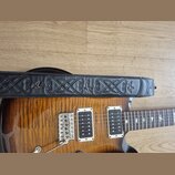 Detail elektrické kytary s vyznačenou dřevěnou strukturou a dvěma humbuckery v bílých rámečcích. Přes tělo kytary je položen černý kožený popruh s vy…