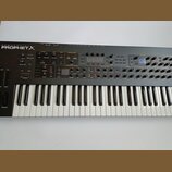 Černý syntetizátor s klaviaturou a ovládacím panelem s mnoha knoflíky, tlačítky a displeji. V levém horním rohu je bílý nápis SEQUENTIAL PROPHET X. P…