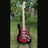 Elektrická kytara typu Stratocaster s trupem v červeně-černém sunburst provedení a hnědým želvovinovým pickguardem. Na hlavě hmatníku je nápis Squier…