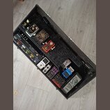 Otevřený černý transportní kufr s vnitřním pedalboardem, na kterém je umístěno několik efektů. V horní části jsou pedály s nápisy IONIK, Deluxe a v č…