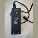 Černý obdélníkový adaptér s nápisy HD 2 a iRig. Zařízení má připojený USB kabel a jeden volný konektor Lightning. V dolní části jsou vyznačené ikony…