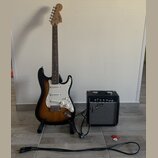 Elektrická kytara s trupem v barevnosti sunburst, bílým pickguardem a hnedým hmatníkem. Na hlavě kytary je nápis Fender. Vedle kytary stojí černý kom…