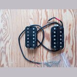 Dvě černé humbucker snímače s kovovými šroubovnicemi, připojené k sobě černým kabelem s odhalenými vodiči na koncích. Na obou snímačích je svisle nap…