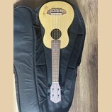 Světlý dřevěný ukulele položené na černém obalu. Na horní části korpusu je vidět hnědé logo s nápisem, který není plně čitelný. Na hlavě nástroje je…
