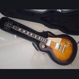Elektrická kytara typu Les Paul v barvě sunburst s tmavě hnědým okrajem a žlutým středem, umístěná v černém tvrdém kufru. Na hlavě hmatníku je nápis…