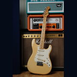 Elektrická kytara typu Stratocaster v krémové barvě s bílým pickguardem a světle hnědým javorovým hmatníkem. Na hlavě kytary je viditelné logo Fender…
