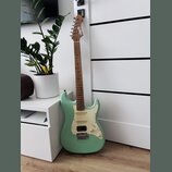 Světle zelená elektrická kytara typu Stratocaster s bílým pickguardem a hnědým hmatníkem. Na hlavě nástroje je bílé logo "Squier". Kytara má konfigur…