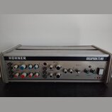 Stříbrný elektronický nástroj s kovovým šasi a horní mřížkou. Na předním panelu je vlevo nápis HOHNER a vpravo ORGAPHON 75 MH. Přístroj obsahuje něko…