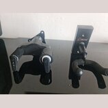 Dvě černé držáky na hudební nástroje z kovu a plastu. Pravý držák má oranžovo-bílé logo s nápisem ORTEGA GUITARS - EST 1994. Povrchy jsou matně černé…