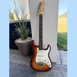 Elektrická kytara s trupem v červenohnědém sunburst provedení a bílým pickguardem. Na hlavě hmatníku je viditelné logo Fender a nápis STRATOCASTER. H…