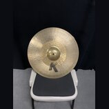 Zlatý kovový činel s kladíčkovaným povrchem, umístěný na bílé židli. V horní části je nápis „Custom Hybrid Ride“ a ve spodní části velké černé logo „…