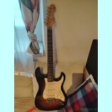Elektrická kytara s trupem v barevném přechodu od černého k hnědému a krčkem z jasného dřeva s tmavým hmatníkem. Na hlavě je viditelné logo Fender. K…