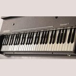 Elektrické piano s šedým, texturovaným povrchem korpusu. Na levé straně je bílý nápis HOHNER a vpravo kurzívou nápis Pianet T. Nástroj má klasickou k…