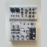Bílá mixovací konzole Yamaha AG06. Na horním panelu jsou vstupy XLR, jacky a konektory RCA s nápisy L/MONO, R, L, R, STEREO OUT a MONITOR OUT. Přední…