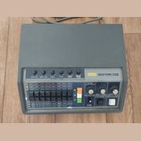 Šedý rytmus stroj s nápisem KORG RHYTHM 55B. Zařízení má ovládací panel s posuvníky pro Bass Drum, Snare Drum, Cymbal, Hi-Hat a TomTom Conga, řadu tl…