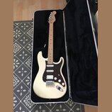 Elektrická kytara s krémovým trupem a hnědým javorovým hmatníkem v černém tvrdém kufru. Nástroj má tmavě červený pickguard, tři snímače a bílé ovláda…