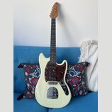 Elektrická kytara s krémovým trupem a hnědým hmatníkem. Na hlavě je nápis "by Taus". Kytara má tmavě červený pickguard s texturou želvy, tři snímače…