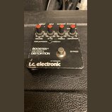 Černý efekt pedál s nápisy BOOSTER+ LINE DRIVER & DISTORTION a logem t.c. electronic OF DENMARK. Horní panel obsahuje čtyři červené oválné knoflíky s…