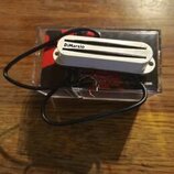 Bílé humbucker snímač s kovovým krytem a černým kabelem. Na krytu je černý nápis DiMarzio. Snímač leží na průhledném plastovém obalu s červeným a čer…