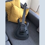 Černá elektrická kytara s matným povrchem a šesti strunami, opřená o žluté polštáře na šedém gauči. Na hlavě hmatníku je bílý nápis STRAT. Kytara má…