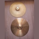 Dvě mosazné činely položené na dřevěné podlaze. Horní činel má v horní části nápis TOP NEW BEAT HI HAT. Oba činely mají jemné kruhové rýhování a vypa…