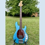 Modrá elektrická basová kytara s čtyřstrunným hmatníkem z tmavého dřeva a naturální hlavou. Tělo má v oblasti snímačů tmavý červený pickguard s textu…