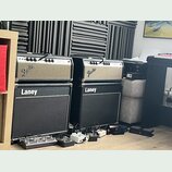 Dvě kytarové komba značky Fender umístěná na dvou černých kytarových boxech značky Laney. Komba mají stříbrný přední panel s nápisy Fender a v pravém…