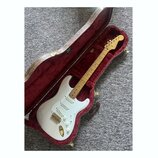 Bílá elektrická kytara typu Stratocaster s javorovým hmatníkem a zlatým hardwarem v otevřeném hnědém tvrdém kufru s červeným polstrováním. Na hlavě k…