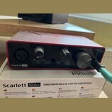 Červená USB audio rozhraní s nápisy Scarlett Solo a Focusrite. Na předním panelu jsou dva ovratové knofíky, jeden velký kruhový knofík MONITOR a vstu…
