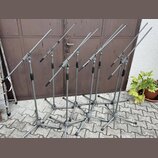 Šest šedých mikrofonních stojanů s boom rameny umístěných na dlažbové ploše před hnědými dveřmi. Stojany jsou vyrobeny z kovu s černými plastovými ut…