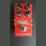 Červený efekt pedál s nápisy tc electronic, HALL OF FAME 2 a REVERB. Obsahuje čtyři černé oválné ovládací knoflíky, jeden stříbrný kovový spínač a ma…