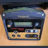 Černý trigger modul značky Roland, model TM-2. Zařízení má v horní části konektory s nápisy L/MONO OUTPUT, R OUTPUT, 1 TRIG IN, 2 TRIG IN a DC IN PSD…