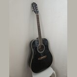 Černá akustická kytara s bílým lemováním korpusu a hnědým hmatníkem. Na hlavě je bílý nápis Epiphone. Nástroj je opřen o světlou stěnu a stojí na bíl…