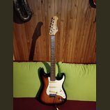 Elektrická kytara s trupem v barevném přechodu od černé přes červenou k žluté a bílým pickguardem. Na hlavě hmatníku je vidět nápis STRATOCASTER. Nás…