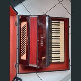 Červená akordeon v černém kufru s červeným vnitřkem. Na pravé straně je klávesnice a na levé tlačítka s nápisem TANGO II S. Na středním červeném kryt…