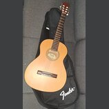 Akustická kytara s přírodním dřevěným povrchem těla a tmavým hmatníkem. Na úzkém pásmu v rosetě je nápis Fender. Kytara leží na černém obalu s bílým…