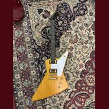 Elektrická kytara s tělem ve tvaru Explorer v přirozené dřevěné barvě a bílým pickguardem. Na hlavě hmatníku je viditelné logo Gibson. Nástroj má dva…