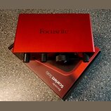 Červený audio rozhraní s nápisem Focusrite na horní straně. Na předním panelu jsou čtyři černé ovladače a nápis Scarlett Solo. Zařízení leží na červe…