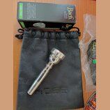 Stříbrný moutur z kovu položený na černém koženém vak s vyraženým nápisem KOSS. Nad nimi je černá krabička s bílými nápisy Bach Genuine Mouthpiece a…