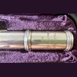 Detail stříbrného kovového dílu flétny v fialovém polstrování pouzdra. Na povrchu je vyryto TAKUMI, NISHIMA FLUTE COLL. a JAPAN. Povrch vykazuje drob…