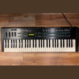 Černý digitální syntetizátor s bílými a černými klávesami. Na horním panelu je nápis YAMAHA DX11 a malý zelený displej s textem PLAY-SINGLE 004: Dim…