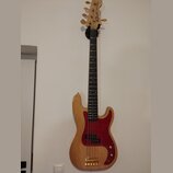 Světle hnědá elektrická basová kytara s přírodním dřevěným povrchem a červeným pickguardem. Na hlavě hmatníku je nápis Fender PRECISION. Nástroj má z…