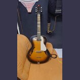 Polodupatá elektrická kytara s korpusem v přechodové barvě od žluté po černou. Nástroj má bílý plechový kryt (pickguard) a bílé ladičky na hlavě. Na…