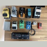 Sestava efektů pro kytaru rozložená na dřevěném povrchu. V horní řadě jsou pedály Behringer GRAPHIC EQUALIZER EQ700, AMPLITUBE, TONEX, S-BAND a BOSS…