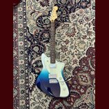 Elektrická kytara s trupem v modrém přechodovém odstínu (fade) od tmavě modré po světle modrou. Na hlavě hmatníku je viditelné logo Fender. Nástroj m…