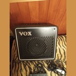 Černý kytarový kombo zesilovač s bílým rámečkem a mřížkou z kovového pletiva. Na přední straně je umístěno stříbrné logo VOX. Před přístrojem leží če…
